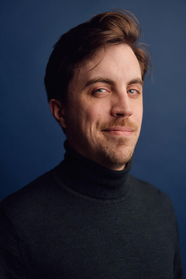 Headshot by Olivia Spencer Felix Trench wearing a dark green rollneck jumper against a blue background.