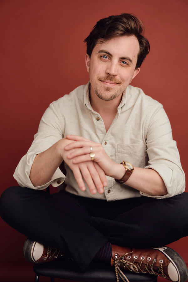 Headshot by Olivia Spencer Felix Trench sitting cross legged against a dark red background